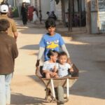 Children playing outdoors, one pushing another in a wheelbarrow.