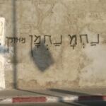 Graffiti in Hebrew on a weathered wall with faded paint and a shadow stain.