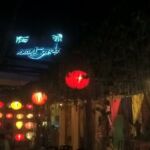 A nighttime street scene illuminated by hanging lanterns and a glowing blue sign.